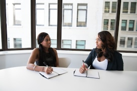 Two individuals are sitting at a white table in a bright room with large windows. They are facing each other, engaged in conversation, while taking notes in notebooks. Both appear attentive and focused.