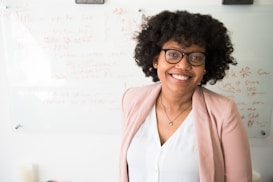 A person with curly hair and glasses is smiling while standing in front of a whiteboard filled with written notes and diagrams. They are wearing a light pink blazer and a white top, conveying a professional setting.