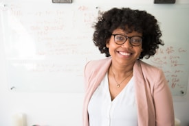 A person with curly hair and glasses is smiling while standing in front of a whiteboard filled with written notes and diagrams. They are wearing a light pink blazer and a white top, conveying a professional setting.