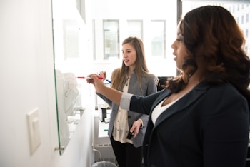 Two women standing in an office environment, engaged in a collaborative work session at a whiteboard. One woman is writing with a red marker while the other observes. Both are dressed in professional attire.