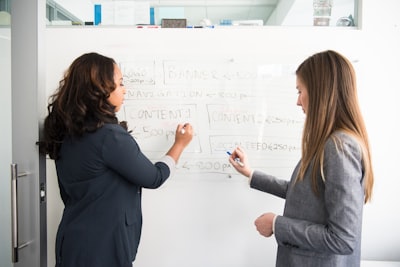 two women writing on whiteboard varied teams background