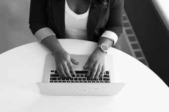 A person typing on a laptop while sitting at a round white table. They are wearing a dark jacket with rolled-up sleeves and a watch on their left wrist.