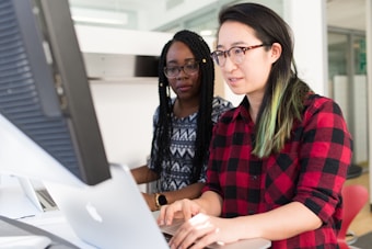 Two people are working together on a project at a desk. One person is wearing glasses and a red plaid shirt, and the other has long braids and a patterned shirt. They are focused on the computer screens in front of them in a modern, well-lit office space.