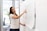 A woman is standing in front of a large sheet of paper attached to a wall, which contains sketches of a website layout. She is pointing to a specific section on the paper, seemingly explaining or presenting the design. The setting appears to be an office with glass walls and plants.