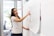 A woman is standing in front of a large sheet of paper attached to a wall, which contains sketches of a website layout. She is pointing to a specific section on the paper, seemingly explaining or presenting the design. The setting appears to be an office with glass walls and plants.