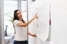 A woman is standing in front of a large sheet of paper attached to a wall, which contains sketches of a website layout. She is pointing to a specific section on the paper, seemingly explaining or presenting the design. The setting appears to be an office with glass walls and plants.