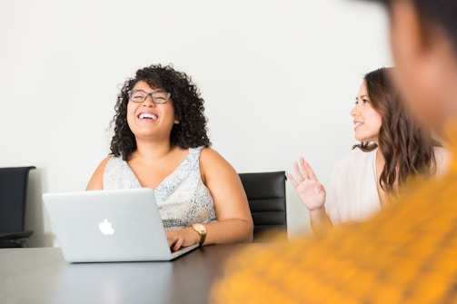 Two people are seated at a table in a bright room. One person is using a laptop and laughing, while the other is speaking with a friendly expression. They appear to be engaged in a lively conversation.