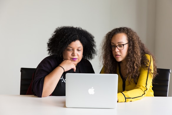 Two people are sitting at a table, focused on a laptop screen. One person has dark curly hair and is wearing a purple shirt, while the other has long curly hair and is wearing glasses and a yellow shirt. They appear to be engaged in a collaborative activity or discussion.