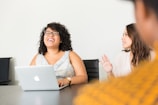 A woman is sitting at a conference table with a laptop in front of her, smiling and laughing. Another woman is sitting next to her, appearing engaged in the conversation. The setting is a meeting or casual office environment, with a focus on collaboration and interaction.