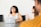 A woman is sitting at a conference table with a laptop in front of her, smiling and laughing. Another woman is sitting next to her, appearing engaged in the conversation. The setting is a meeting or casual office environment, with a focus on collaboration and interaction.