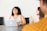 A woman is sitting at a conference table with a laptop in front of her, smiling and laughing. Another woman is sitting next to her, appearing engaged in the conversation. The setting is a meeting or casual office environment, with a focus on collaboration and interaction.