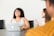 A woman is sitting at a conference table with a laptop in front of her, smiling and laughing. Another woman is sitting next to her, appearing engaged in the conversation. The setting is a meeting or casual office environment, with a focus on collaboration and interaction.