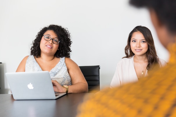 Two women are sitting at a table, engaged in a conversation. One woman is using a laptop placed in front of her. Both appear attentive and focused.