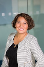 A person with curly short hair is smiling, wearing a striped blazer over a black top and a gold necklace. The background is an office-like setting with a glass partition.
