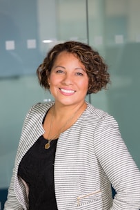 A person with curly short hair is smiling, wearing a striped blazer over a black top and a gold necklace. The background is an office-like setting with a glass partition.