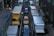 A congested urban street with various delivery trucks and a yellow taxi in separate lanes. Large vehicles, including Penske and U-Haul trucks, dominate the scene, surrounded by tall buildings. Pedestrians are visible on the sidewalks, adding to the bustling city atmosphere.