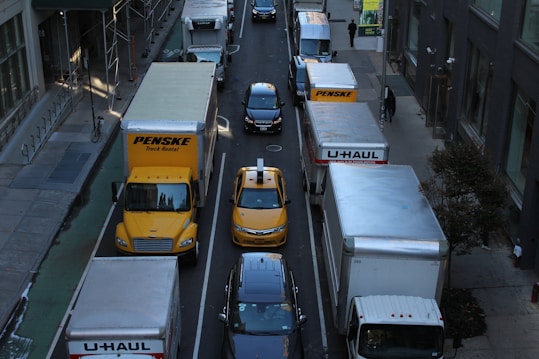A congested urban street with various delivery trucks and a yellow taxi in separate lanes. Large vehicles, including Penske and U-Haul trucks, dominate the scene, surrounded by tall buildings. Pedestrians are visible on the sidewalks, adding to the bustling city atmosphere.