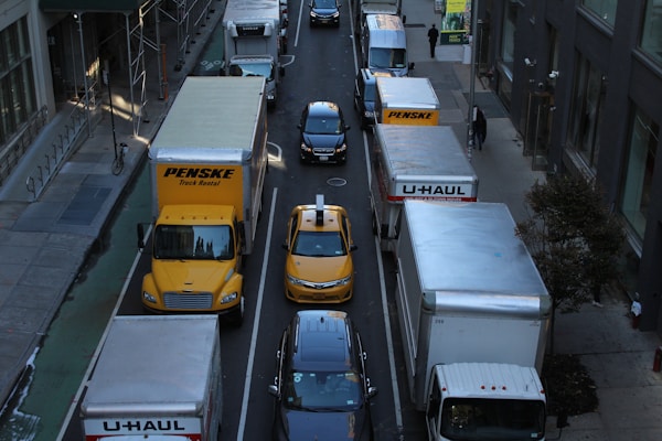 A congested urban street with various delivery trucks and a yellow taxi in separate lanes. Large vehicles, including Penske and U-Haul trucks, dominate the scene, surrounded by tall buildings. Pedestrians are visible on the sidewalks, adding to the bustling city atmosphere.
