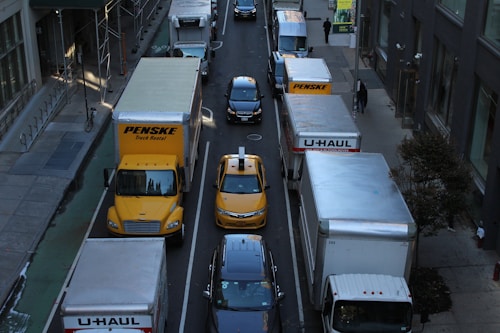 A congested urban street with various delivery trucks and a yellow taxi in separate lanes. Large vehicles, including Penske and U-Haul trucks, dominate the scene, surrounded by tall buildings. Pedestrians are visible on the sidewalks, adding to the bustling city atmosphere.
