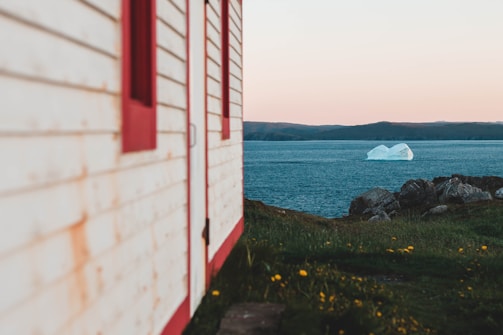 A white and red wooden building is partially visible in the foreground on the left, with a vast ocean extending into the distance. An iceberg floats on the water near the horizon. The landscape includes rocky terrain and green grass dotted with small yellow flowers. The sky transitions from light blue to a soft orange closer to the horizon.