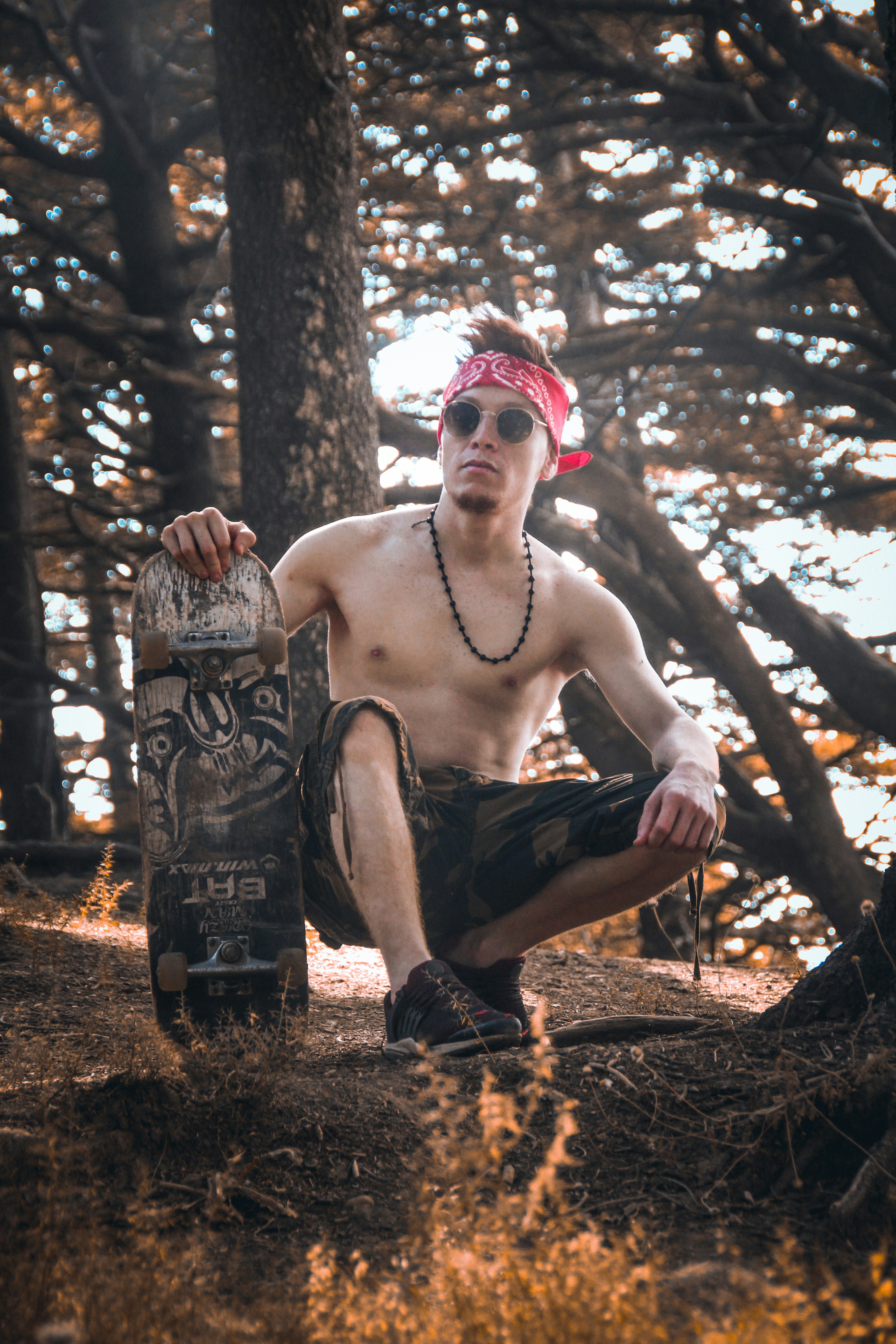 man squatting and holding skateboard near trees