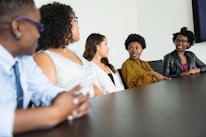 A group of five people sit around a dark wood conference table in discussion. They appear to be engaged and attentive, with one individual speaking and others listening intently. The group includes both men and women dressed in business casual attire. The background is a plain, light-colored wall.