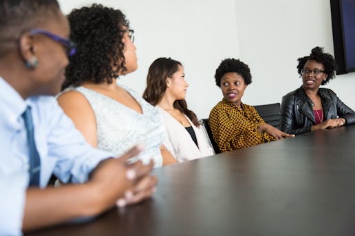 A group of five people sit around a dark wood conference table in discussion. They appear to be engaged and attentive, with one individual speaking and others listening intently. The group includes both men and women dressed in business casual attire. The background is a plain, light-colored wall.
