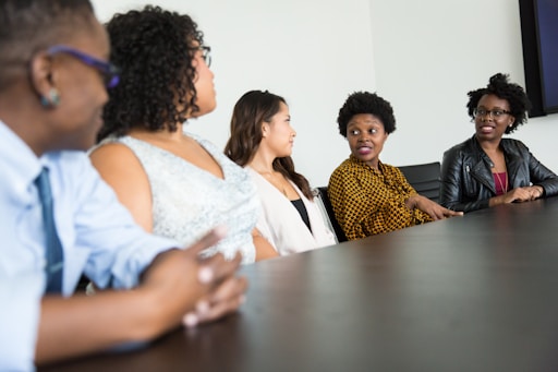 A group of five people sit around a dark wood conference table in discussion. They appear to be engaged and attentive, with one individual speaking and others listening intently. The group includes both men and women dressed in business casual attire. The background is a plain, light-colored wall.