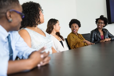 A group of five people sit around a dark wood conference table in discussion. They appear to be engaged and attentive, with one individual speaking and others listening intently. The group includes both men and women dressed in business casual attire. The background is a plain, light-colored wall.