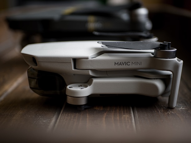 A compact, folded drone labeled 'Mavic Mini' rests on a wooden surface. The focus is on its sleek and modern design, characterized by its light gray body and streamlined construction.