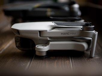 A compact, folded drone labeled 'Mavic Mini' rests on a wooden surface. The focus is on its sleek and modern design, characterized by its light gray body and streamlined construction.