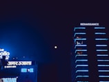 A nighttime urban scene featuring a tall building with neon blue lights that trace its edges, identified by the sign 'RENAISSANCE' at the top. Adjacent to it are structures with various illuminated signs, including 'INOX' and several signs in a non-Latin script, all highlighted against a dark sky with a visible moon.