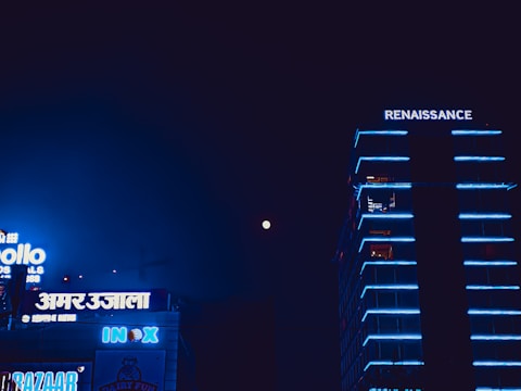 A nighttime urban scene featuring a tall building with neon blue lights that trace its edges, identified by the sign 'RENAISSANCE' at the top. Adjacent to it are structures with various illuminated signs, including 'INOX' and several signs in a non-Latin script, all highlighted against a dark sky with a visible moon.