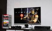 A flat-screen TV displays a video game interface featuring a character from Mortal Kombat in the center. Beside the TV is a stack of video game cases for the PlayStation 3, including titles like FIFA 14 and Grand Theft Auto. Below the TV, there is a gaming console, controllers, and other gaming equipment.