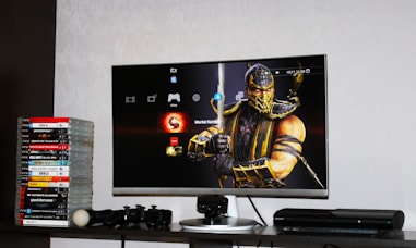 A flat-screen TV displays a video game interface featuring a character from Mortal Kombat in the center. Beside the TV is a stack of video game cases for the PlayStation 3, including titles like FIFA 14 and Grand Theft Auto. Below the TV, there is a gaming console, controllers, and other gaming equipment.