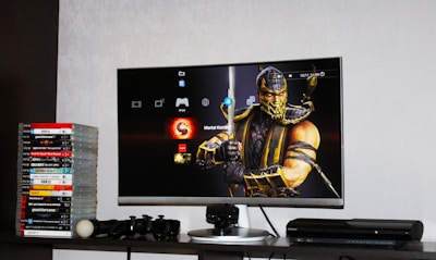 A flat-screen TV displays a video game interface featuring a character from Mortal Kombat in the center. Beside the TV is a stack of video game cases for the PlayStation 3, including titles like FIFA 14 and Grand Theft Auto. Below the TV, there is a gaming console, controllers, and other gaming equipment.