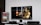 A flat-screen TV displays a video game interface featuring a character from Mortal Kombat in the center. Beside the TV is a stack of video game cases for the PlayStation 3, including titles like FIFA 14 and Grand Theft Auto. Below the TV, there is a gaming console, controllers, and other gaming equipment.