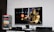 A flat-screen TV displays a video game interface featuring a character from Mortal Kombat in the center. Beside the TV is a stack of video game cases for the PlayStation 3, including titles like FIFA 14 and Grand Theft Auto. Below the TV, there is a gaming console, controllers, and other gaming equipment.