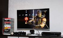 A flat-screen TV displays a video game interface featuring a character from Mortal Kombat in the center. Beside the TV is a stack of video game cases for the PlayStation 3, including titles like FIFA 14 and Grand Theft Auto. Below the TV, there is a gaming console, controllers, and other gaming equipment.