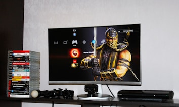 A flat-screen TV displays a video game interface featuring a character from Mortal Kombat in the center. Beside the TV is a stack of video game cases for the PlayStation 3, including titles like FIFA 14 and Grand Theft Auto. Below the TV, there is a gaming console, controllers, and other gaming equipment.