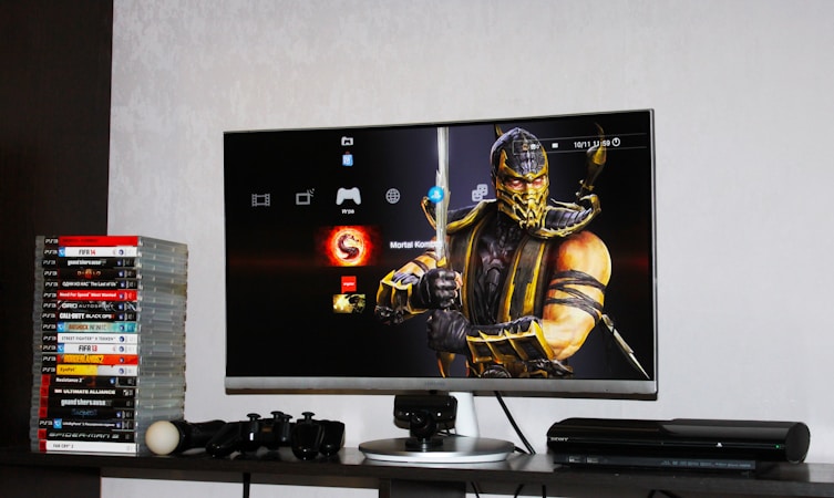 A flat-screen TV displays a video game interface featuring a character from Mortal Kombat in the center. Beside the TV is a stack of video game cases for the PlayStation 3, including titles like FIFA 14 and Grand Theft Auto. Below the TV, there is a gaming console, controllers, and other gaming equipment.