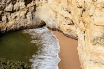 A small beach hidden between large, rocky cliffs with waves gently washing onto the golden sand. The rock walls are rugged and textured, with patches of vegetation visible at the top.