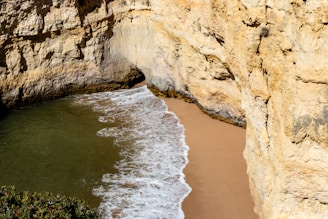 A small beach hidden between large, rocky cliffs with waves gently washing onto the golden sand. The rock walls are rugged and textured, with patches of vegetation visible at the top.