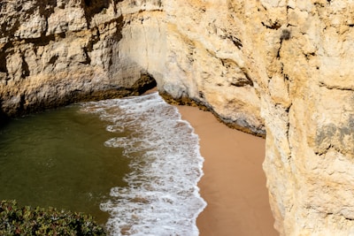 A small beach hidden between large, rocky cliffs with waves gently washing onto the golden sand. The rock walls are rugged and textured, with patches of vegetation visible at the top.