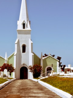 A white church with a tall spire stands against a clear blue sky. The church is flanked by green grass and a pathway leads up to its arched wooden door. Red flowering shrubs are on either side of the entrance, and tombstones are visible in the background.