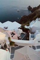 Overlooking a scenic coastal view with whitewashed buildings and terraces descending along a cliffside. In the background, the deep blue sea is visible, with a small boat floating near the rugged coastline. Umbrellas and deck chairs are situated on the terraces, accompanied by a mix of greenery.