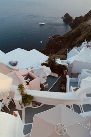 Overlooking a scenic coastal view with whitewashed buildings and terraces descending along a cliffside. In the background, the deep blue sea is visible, with a small boat floating near the rugged coastline. Umbrellas and deck chairs are situated on the terraces, accompanied by a mix of greenery.