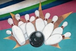 A stylized illustration featuring a black bowling ball striking a set of white bowling pins against a colorful, abstract background with stripes of blue, green, and pink.