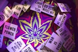 A psychedelic background with vibrant colors frames a series of protest signs. The signs display messages advocating for social justice and rights, such as 'Women's Liberation', 'Flower Power', 'War is Over!', and 'We Shall Overcome'. A large cannabis leaf is prominently centered in the composition.