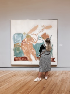 A person with long hair stands in front of an abstract painting in a gallery. The painting features large, expressive brushstrokes in shades of blue, orange, and white. The gallery room has wooden floors and neutral-colored walls, enhancing the focus on the artwork.